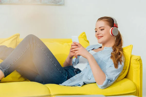 Focus selettivo della donna sorridente in cuffia utilizzando smartphone sul divano in soggiorno — Foto stock