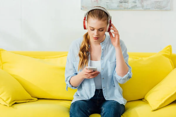 Mulher bonita em fones de ouvido usando smartphone no sofá em casa — Fotografia de Stock