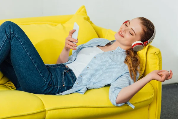 Vista lateral de la chica sonriente escuchando música en los auriculares y utilizando el teléfono inteligente en el sofá - foto de stock