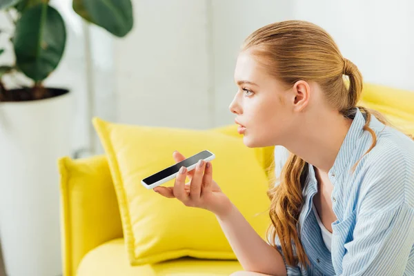 Side view of beautiful girl using speakerphone while talking on smartphone on yellow sofa at home — Stock Photo