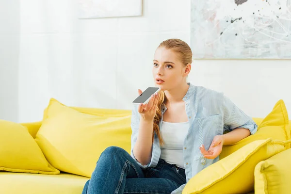 Mujer atractiva usando altavoz mientras habla en el teléfono inteligente en el sofá en casa - foto de stock