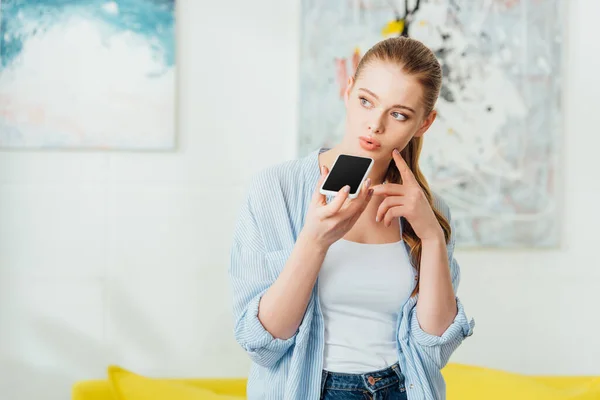 Chica pensativa con el dedo cerca de la mejilla hablando en el teléfono inteligente en casa - foto de stock