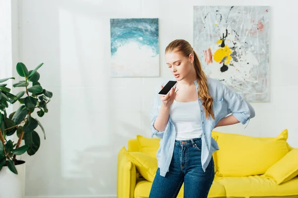 Mulher bonita usando alto-falante enquanto conversa no smartphone na sala de estar — Fotografia de Stock