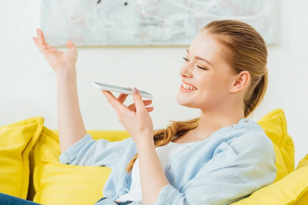 Vista lateral de la mujer sonriente haciendo gestos mientras usa el altavoz en el teléfono inteligente en el sofá - foto de stock
