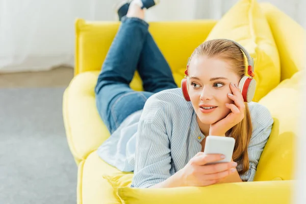 Selective focus of beautiful woman in headphones holding smartphone while lying on couch — Stock Photo