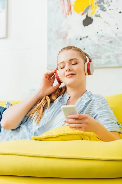Chica atractiva con los ojos cerrados escuchando música en los auriculares y sosteniendo el teléfono inteligente en el sofá - foto de stock