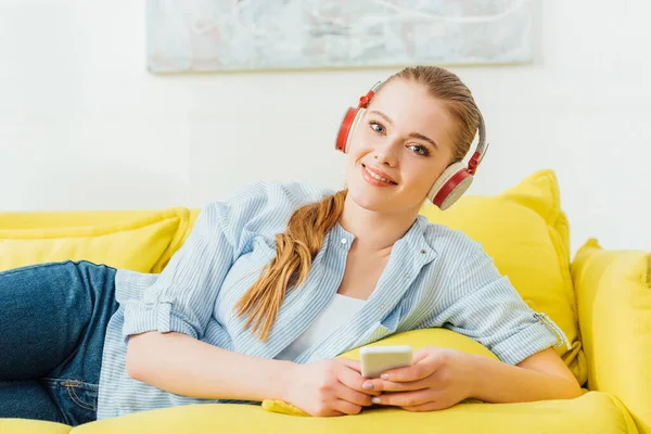 Chica atractiva en auriculares sonriendo a la cámara y sosteniendo el teléfono inteligente en el sofá - foto de stock