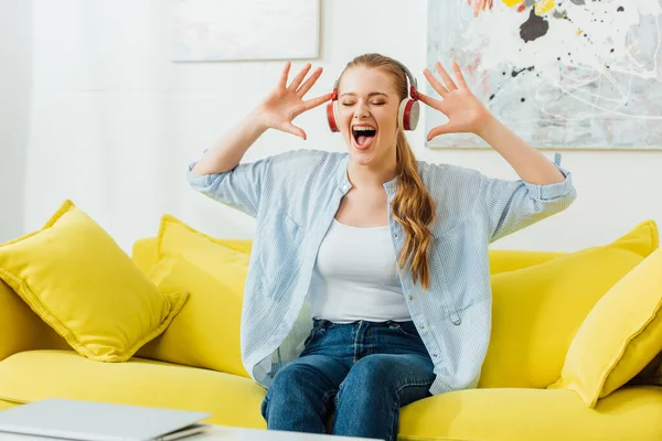 Mulher bonita em fones de ouvido cantando no sofá na sala de estar — Fotografia de Stock