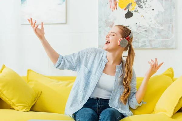 Mulher bonita cantando enquanto ouve música em fones de ouvido no sofá em casa — Fotografia de Stock