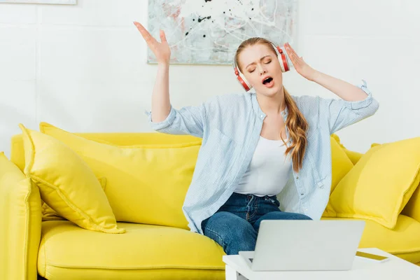 Mulher atraente cantando enquanto ouve música em fones de ouvido no sofá perto de laptop e smartphone na mesa de café — Fotografia de Stock