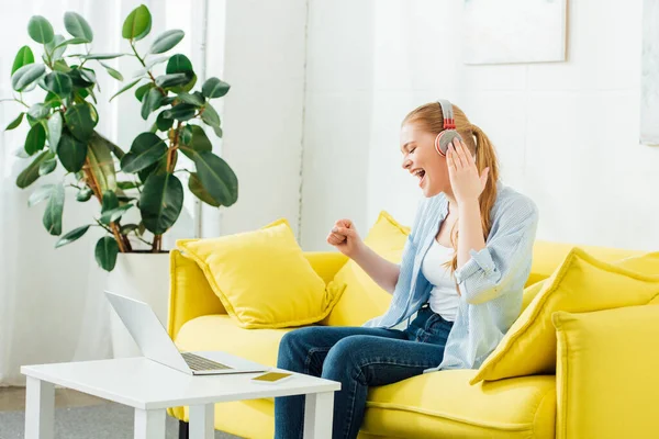 Vista lateral de la chica alegre en los auriculares cantando en el sofá cerca de la computadora portátil y teléfono inteligente en la mesa de café en casa - foto de stock
