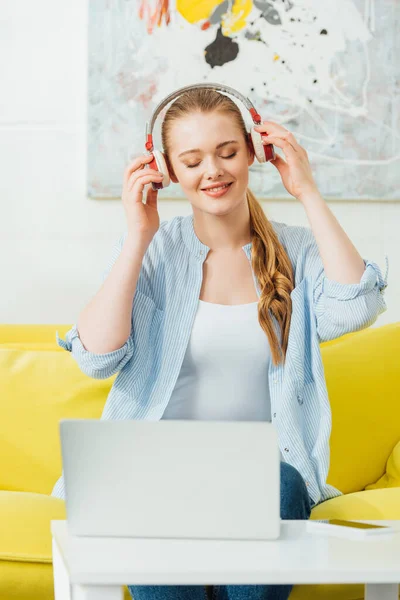 Selective focus of smiling woman listening music in headphones near laptop and smartphone on coffee table in living room — Stock Photo