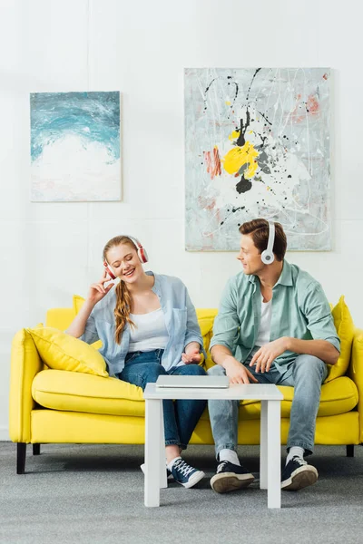 Pareja joven en auriculares sentados en el sofá cerca de la computadora portátil en la mesa de café - foto de stock