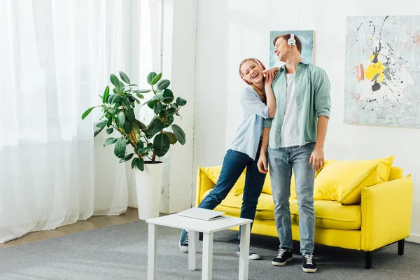 Sorrindo mulher em fones de ouvido abraçando namorado na sala de estar — Fotografia de Stock