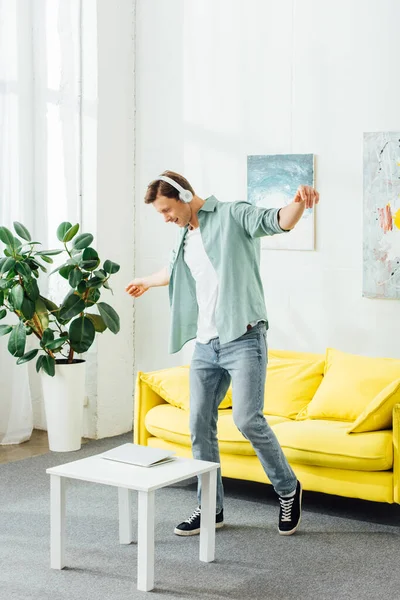 Vista lateral del joven en auriculares bailando cerca de la computadora portátil en la mesa de centro en la sala de estar - foto de stock