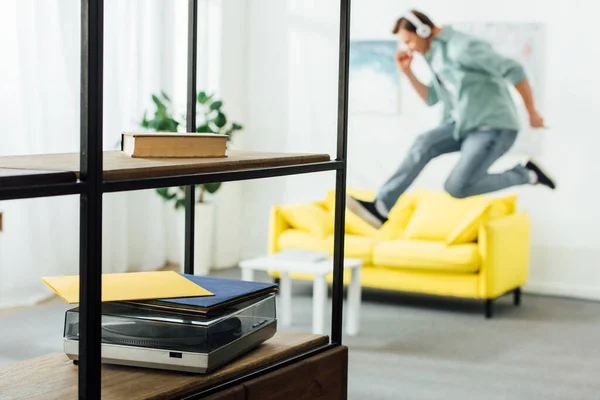 Selective focus of record player and book on shelves of cabinet and man in headphones jumping in living room — Stock Photo