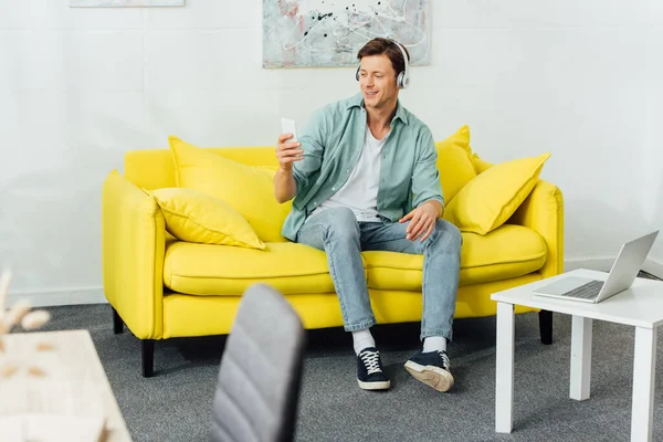 Вибірковий фокус усміхненого чоловіка в навушниках за допомогою смартфона на дивані біля ноутбука на кавовому столі — стокове фото