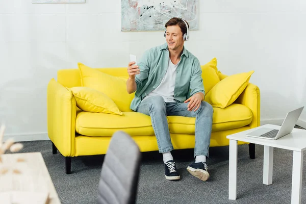 Selective focus of smiling man in headphones using smartphone on couch near laptop on coffee table — Stock Photo