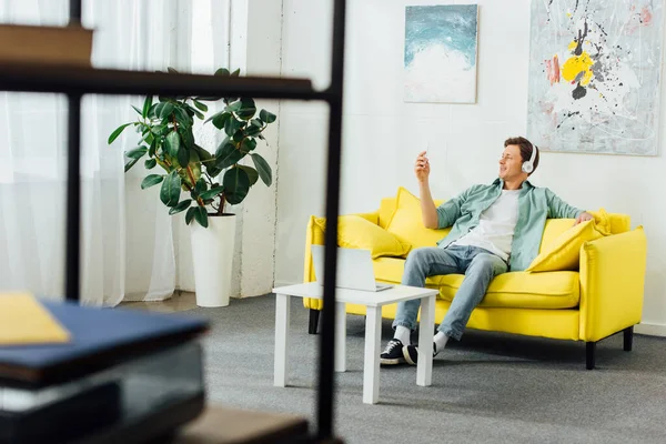 Вибірковий фокус людини в навушниках за допомогою смартфона на дивані біля ноутбука на кавовому столі вдома — стокове фото