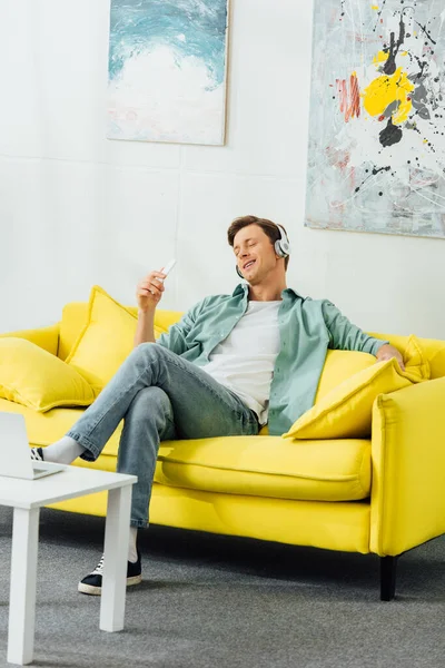 Un bel homme souriant dans un casque tenant un smartphone sur un canapé près d'un ordinateur portable sur une table basse — Photo de stock