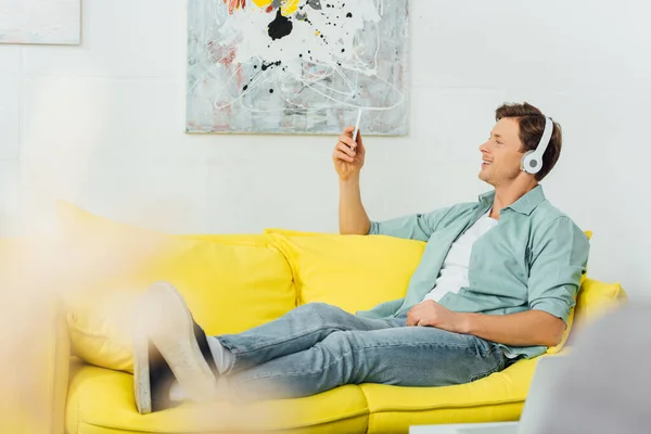 Выборочный фокус улыбающегося мужчины в наушниках с помощью смартфона на диване дома — стоковое фото