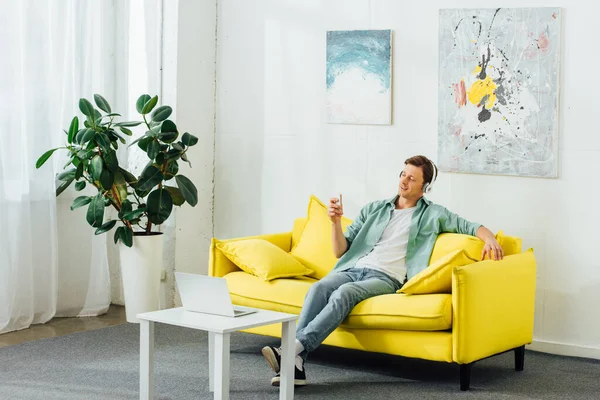 Hombre guapo usando teléfono inteligente mientras escucha música en el sofá cerca de la computadora portátil en la mesa de café - foto de stock