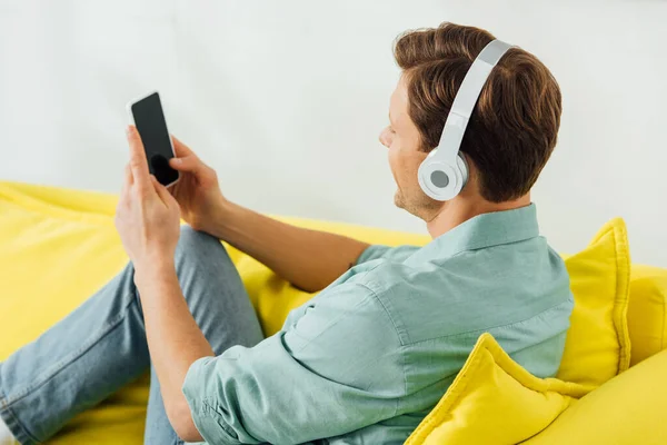 Vista lateral del hombre en auriculares con smartphone en sofá sobre fondo gris - foto de stock