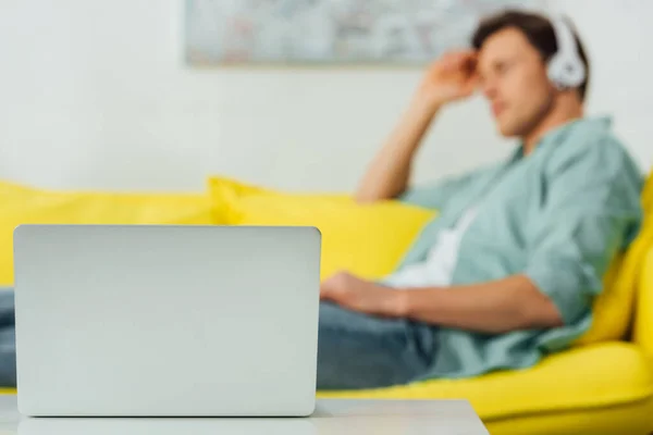 Selective focus of laptop on coffee table and man listening music in headphones on couch — Stock Photo