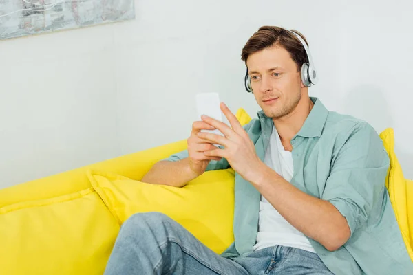 Handsome man in headphones using smartphone on yellow couch at hoe — Stock Photo