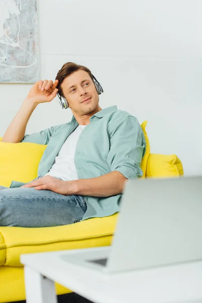 Focus sélectif du bel homme dans les écouteurs regardant loin sur le canapé près de l'ordinateur portable sur la table basse — Photo de stock