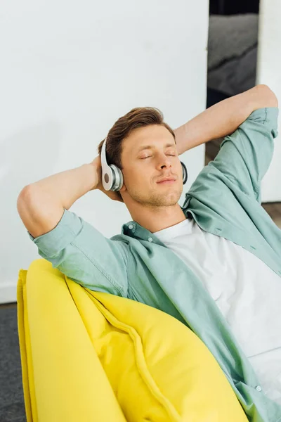 Hombre guapo con los ojos cerrados escuchando música en los auriculares en el sofá en casa - foto de stock