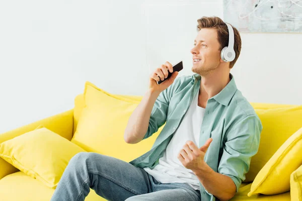 Schöner Mann mit Kopfhörer hält Smartphone zu Hause auf Couch — Stockfoto