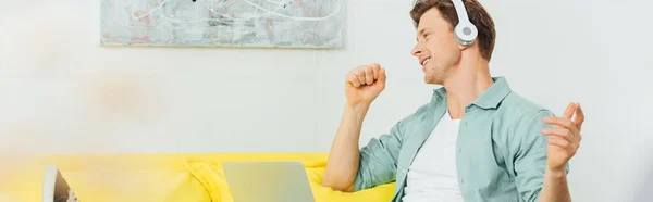 Focus selettivo dell'uomo in cuffia che canta vicino al laptop sul divano, scatto panoramico — Foto stock