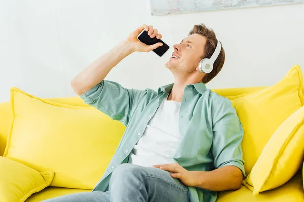 Красивий чоловік у навушниках співає, тримаючи смартфон на жовтому дивані у вітальні — стокове фото