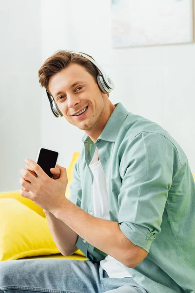 Side view of man in headphones smiling at camera and holding smartphone on couch — Stock Photo