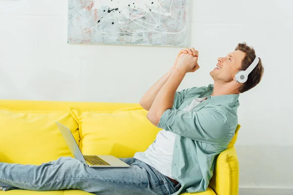 Seitenansicht eines Mannes mit Kopfhörern, der singt, während er Laptop auf Sofa im Wohnzimmer hält — Stockfoto