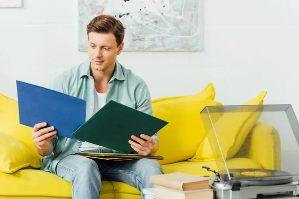 Bonito homem segurando discos de vinil perto do leitor de discos e livros na mesa de café — Fotografia de Stock