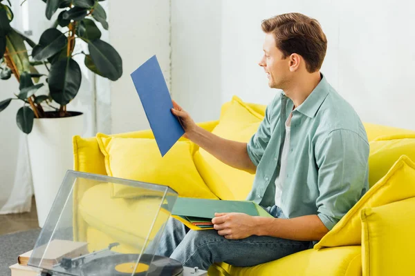 Vista lateral do homem segurando registros de vinil perto de gravador e livros na mesa de café na sala de estar — Fotografia de Stock