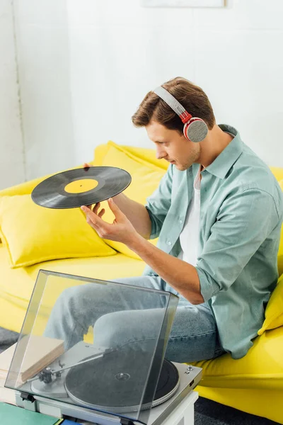 Seitenansicht eines gutaussehenden Mannes mit Kopfhörern, der Schallplatte neben Büchern und Plattenspieler auf dem Couchtisch hält — Stockfoto