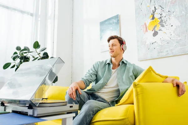 Handsome man in headphones sitting on couch near record player on coffee table — Stock Photo