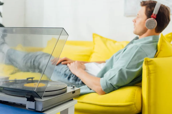 Selective focus of record player on coffee table and man in headphones lying on couch at home — Stock Photo