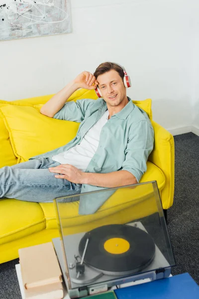 Vista de ángulo alto del tocadiscos y libros en la mesa de café y hombre sonriente en auriculares sentados en el sofá en casa - foto de stock