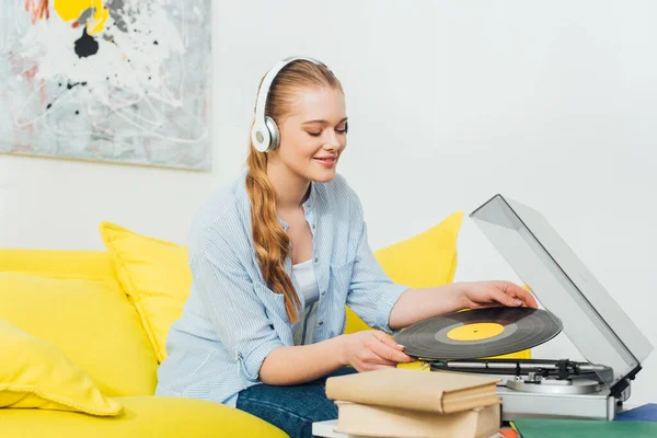 Femme souriante dans des écouteurs tenant un disque vinyle près du tourne-disque et des livres sur la table basse — Photo de stock