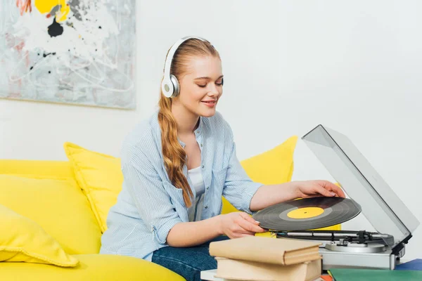 Mulher sorridente em fones de ouvido segurando vinil recorde perto de leitor de discos e livros na mesa de café — Fotografia de Stock