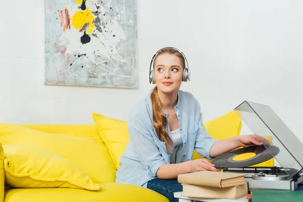 Hermosa mujer en auriculares sosteniendo disco de vinilo cerca de libros y tocadiscos en el sofá en la sala de estar - foto de stock