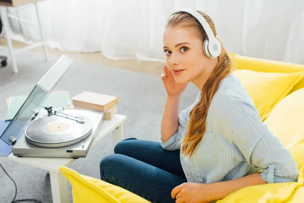 Vista lateral de la mujer mirando a la cámara mientras escucha música en auriculares cerca del reproductor de discos y libros en la mesa de café - foto de stock