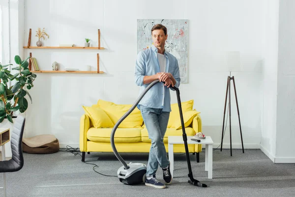 Schöner Mann schaut weg, während er Staubsauger im Wohnzimmer hält — Stockfoto