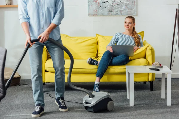 Lächelndes Mädchen mit Laptop schaut Freund mit Staubsauger im Wohnzimmer an — Stockfoto