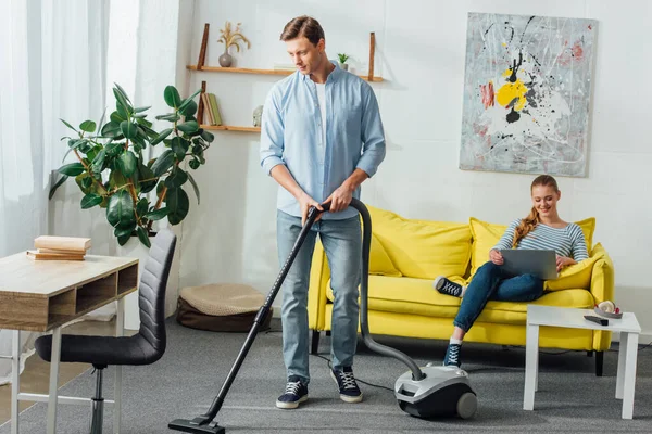 Hombre alfombra de limpieza con aspiradora cerca de la chica sonriente utilizando el ordenador portátil en el sofá en casa - foto de stock