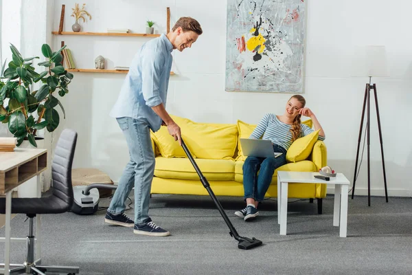 Lächelnder Mann putzt Teppich mit Staubsauger neben Freundin mit Laptop auf Sofa zu Hause — Stockfoto